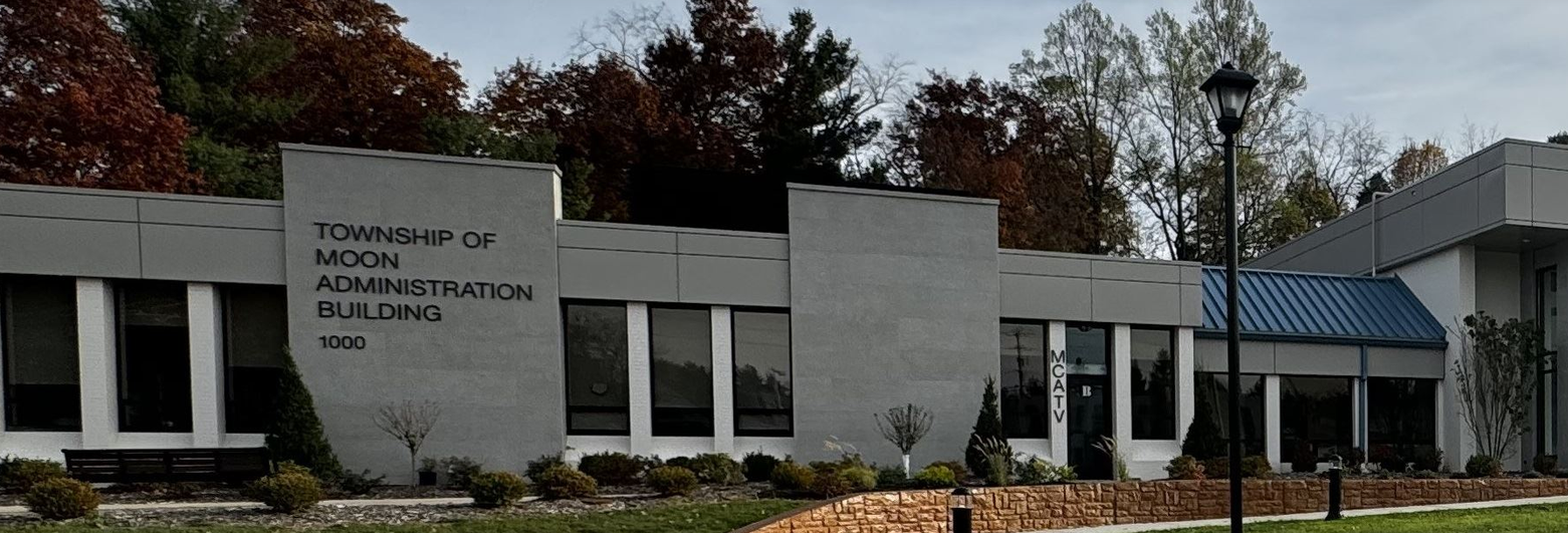 The Moon Township Administration Building, a low grey building with TOWNSHIP OF MOON ADMINISTRATION BUILDING 1000 lettered on the front wall, autumn trees behind it and the MCA-TV entrance to the right.
