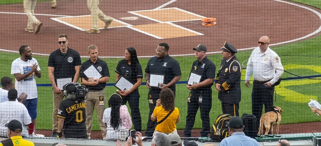 Uniformed officers and other honorees stand on the field at PNC Park holding framed certificates, with police commanders beside them and spectators watching from the stands.